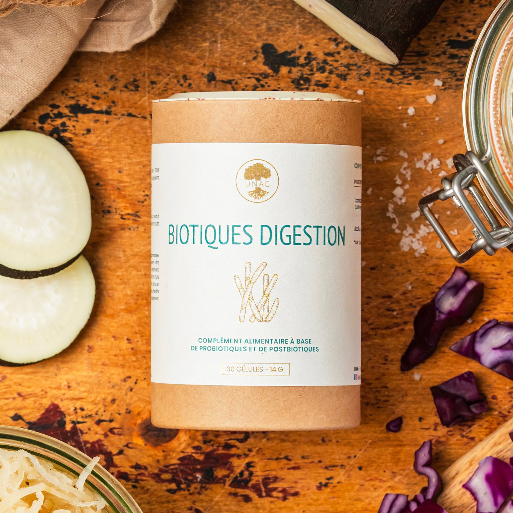Boîte cylindrique en carton du complément alimentaire UNAE Biotiques Digestion posée sur une table en bois, entourée de légumes fermentés, de morceaux de radis noir et de chou rouge.