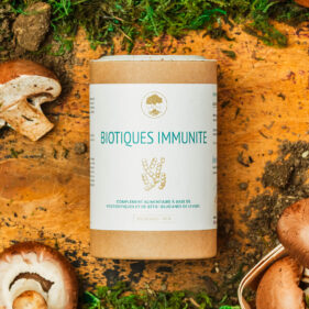 Boîte cylindrique en carton du complément alimentaire UNAE Biotiques Immunité posée sur une surface en bois, entourée de champignons, de mousse verte et de terre.