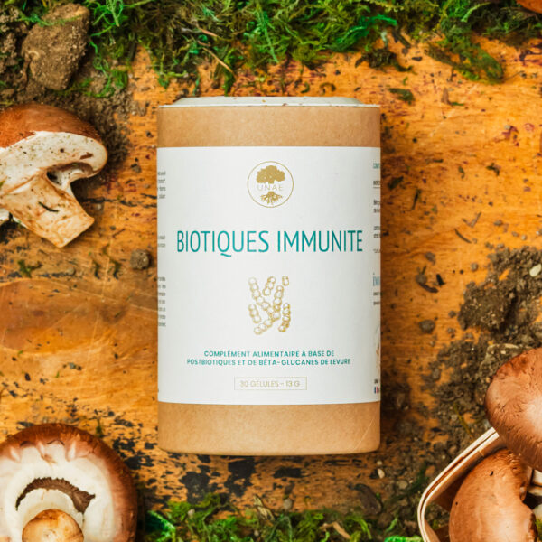 Boîte cylindrique en carton du complément alimentaire UNAE Biotiques Immunité posée sur une surface en bois, entourée de champignons, de mousse verte et de terre.