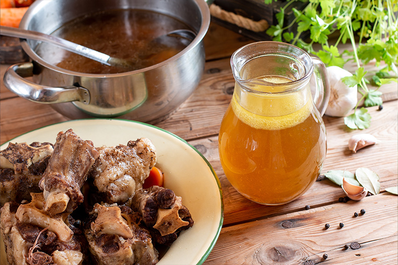 Bouillon d’os maison : casserole remplie de bouillon chaud, pichet de bouillon doré et assiette d’os de bœuf mijotés sur une table en bois, entourés d’herbes fraîches et de gousses d’ail.