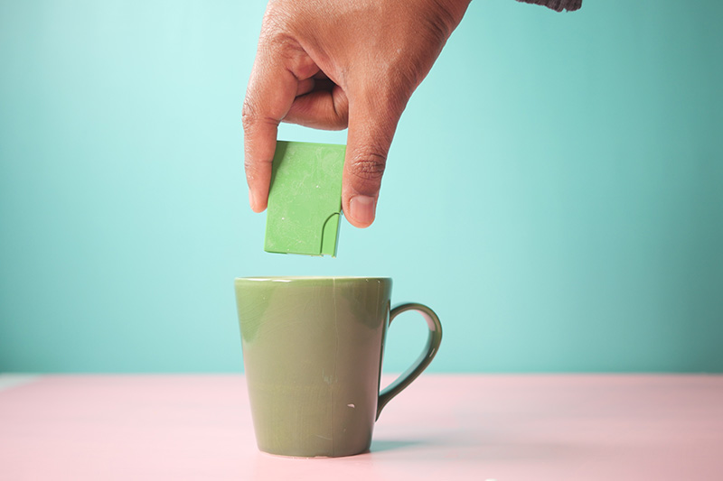 Main tenant du succralose en train d'en ajouter dans une tasse de thé ou café facteur de déséquilibres de notre microbiote dans notre alimentation